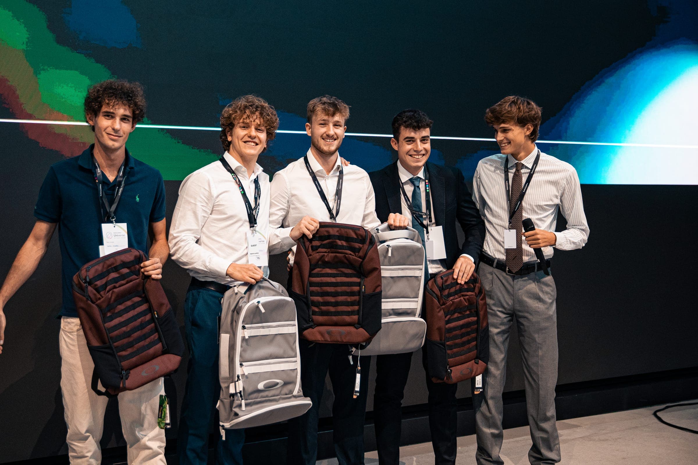 Five young men wearing lanyards smile on stage, each holding a backpack, in front of a large screen showing colourful images
