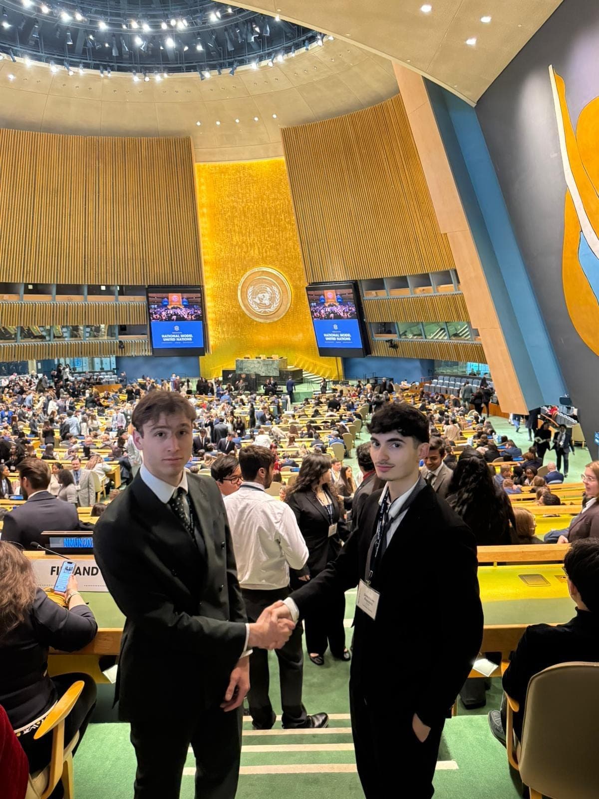 Two young men in dark suits shake hands in a large assembly hall full of students; the screens read National Model United Nations